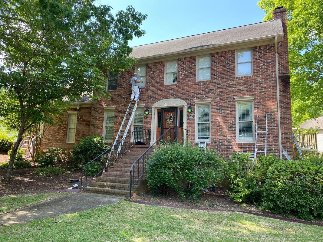 A person on a ladder paints a two-story brick house with a brown roof and green landscaping.