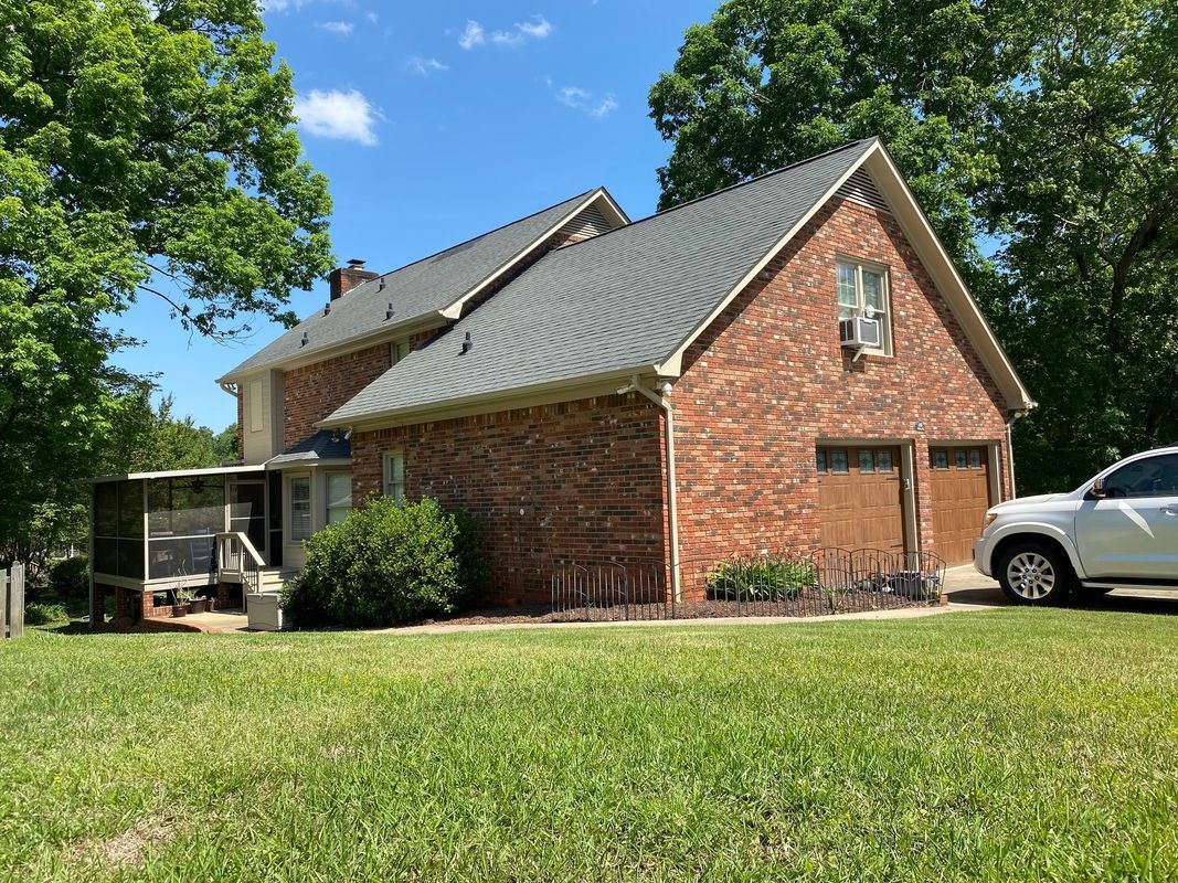 Brick house with garage, screen porch, and car parked on the side, set on a grassy lot under a blue sky.