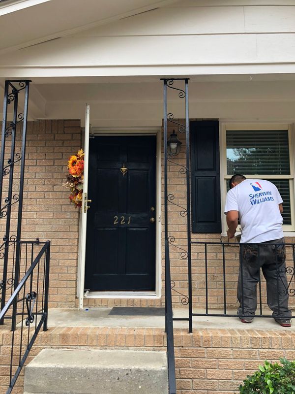A person paints a black railing on a home's porch. The house has a blue door, brick facade, and decorative supports.