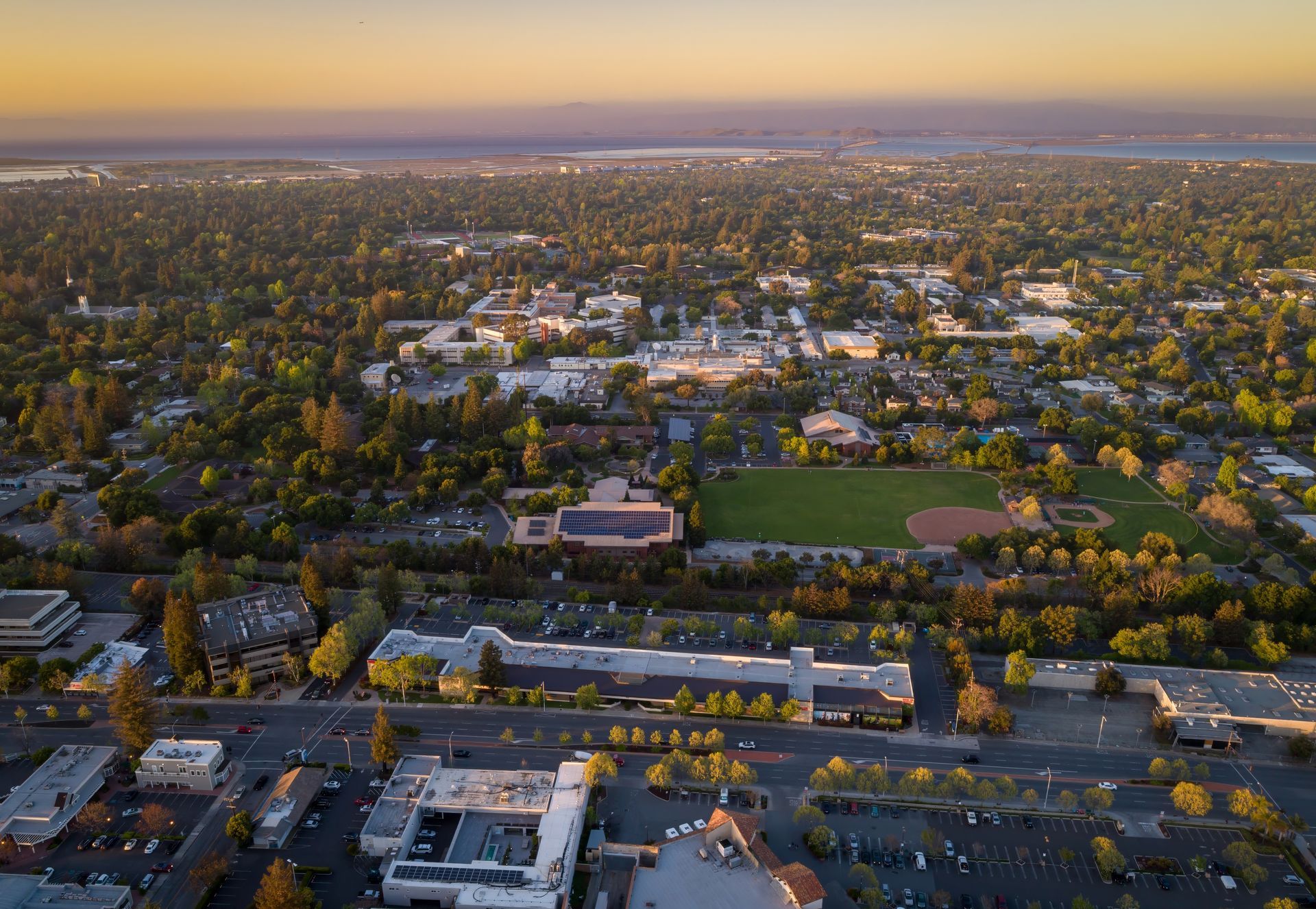 Aerial view of a suburban cityscape at sunset with roads, buildings, and trees stretching to the horizon