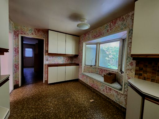 Empty retro kitchen with floral wallpaper, brown patterned floor, white cabinets, and a large window sink area.