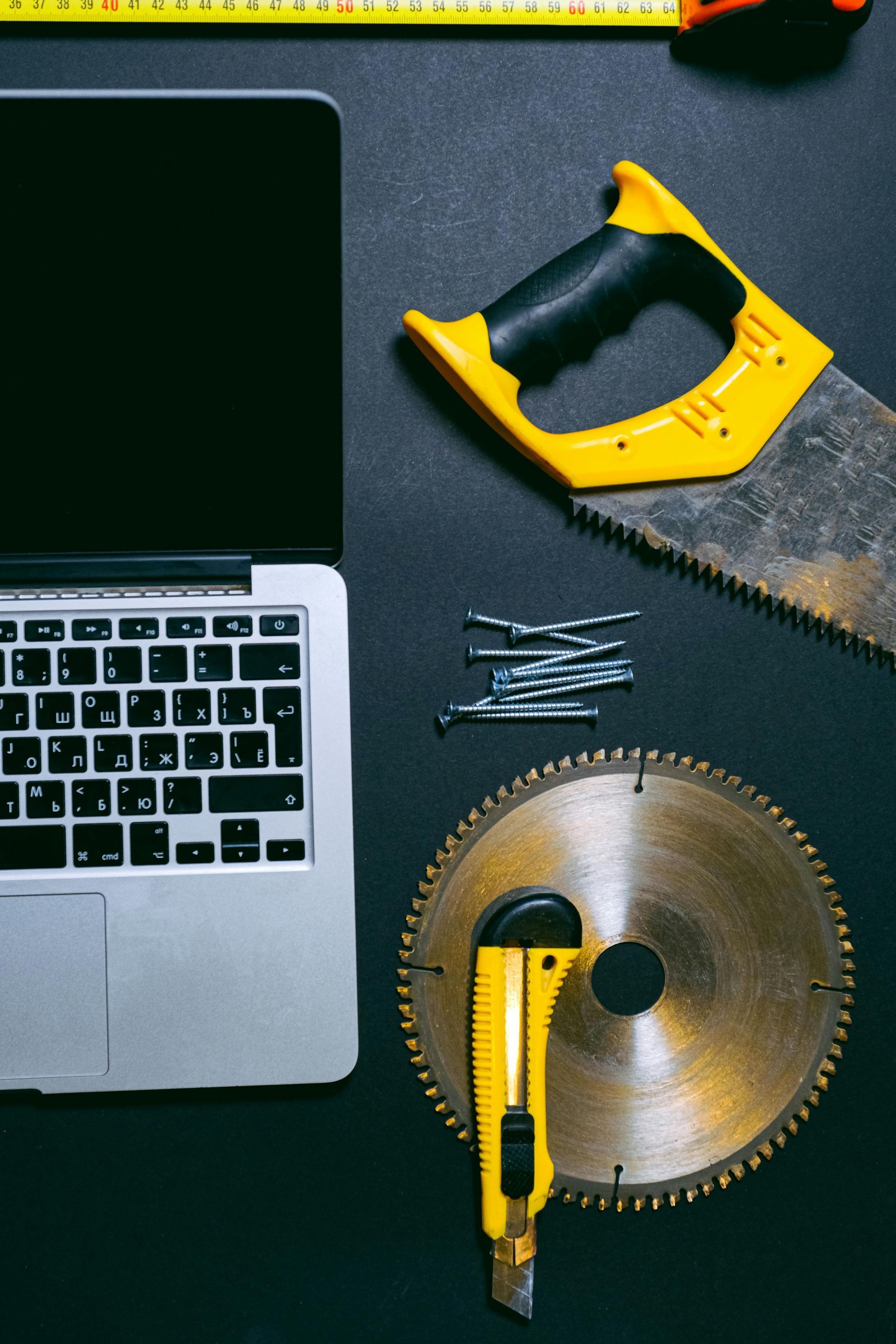 A laptop is surrounded by tools including a circular saw and a tape measure