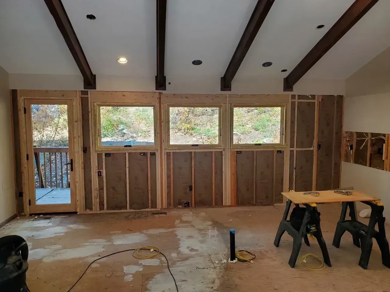 Interior room under construction, with windows and exposed framing. Brown beams on the ceiling, dirty floor, and a sawhorse.