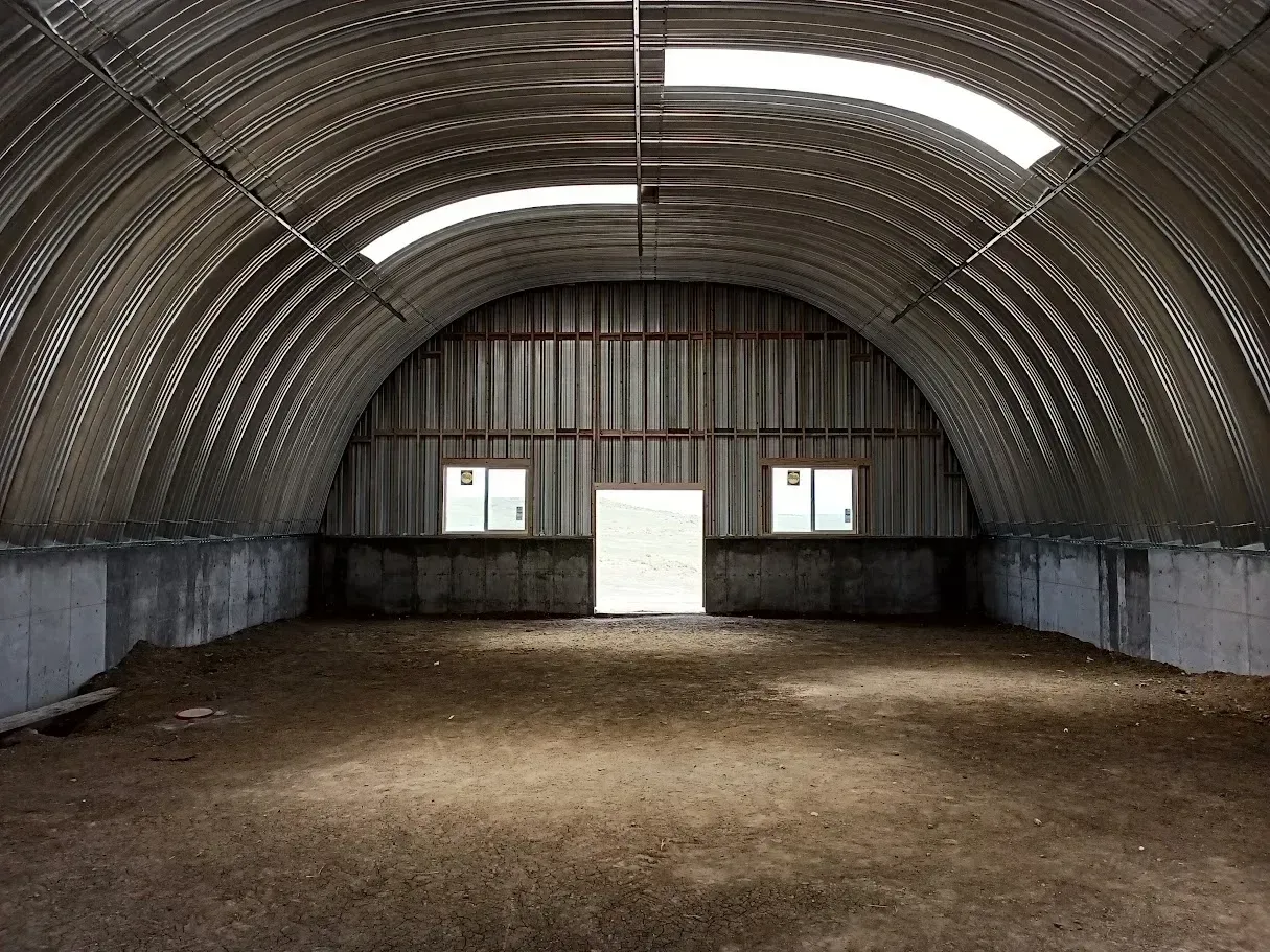 Interior of an empty Quonset hut with a dirt floor, metal arched roof, concrete base, and door with two windows.