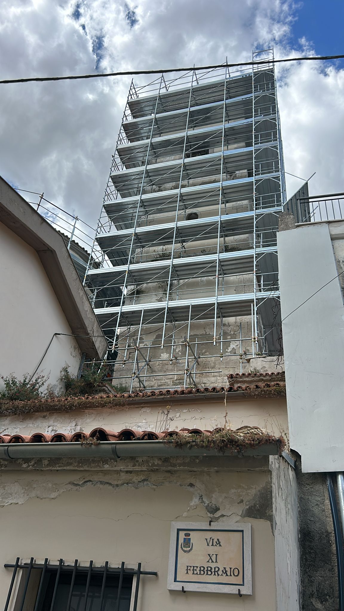 Cantiere: grattacielo incompiuto dietro un edificio basso, impalcature metalliche visibili, cielo nuvoloso.