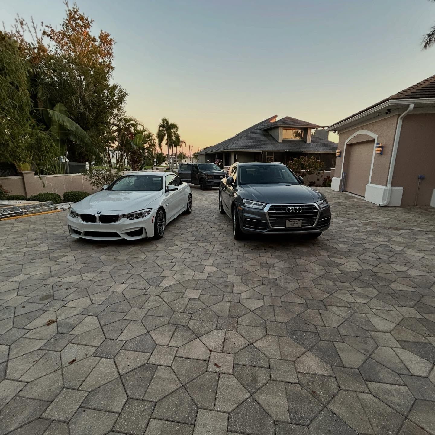 Two parked sedans, white and black, on a stone driveway at sunset, with houses and trees in the background