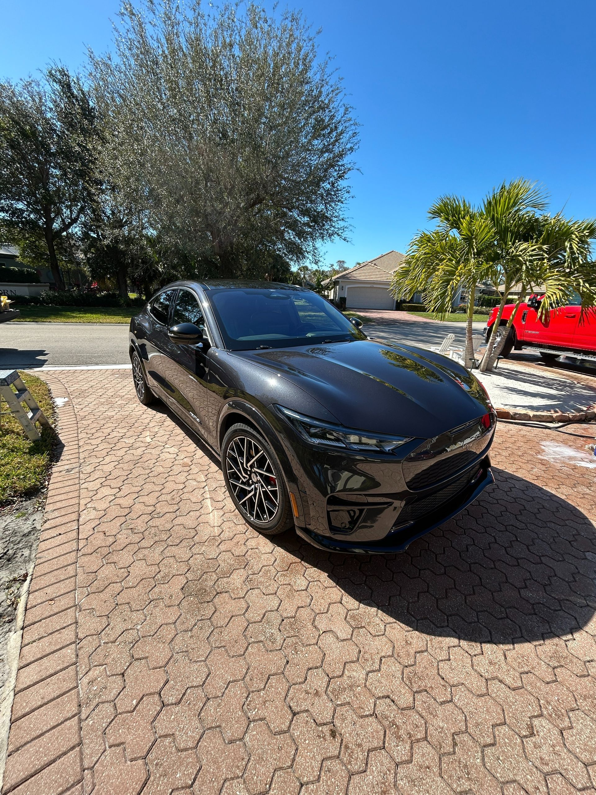 Black SUV parked in a driveway under a sunny sky, with trees and a red vehicle nearby.