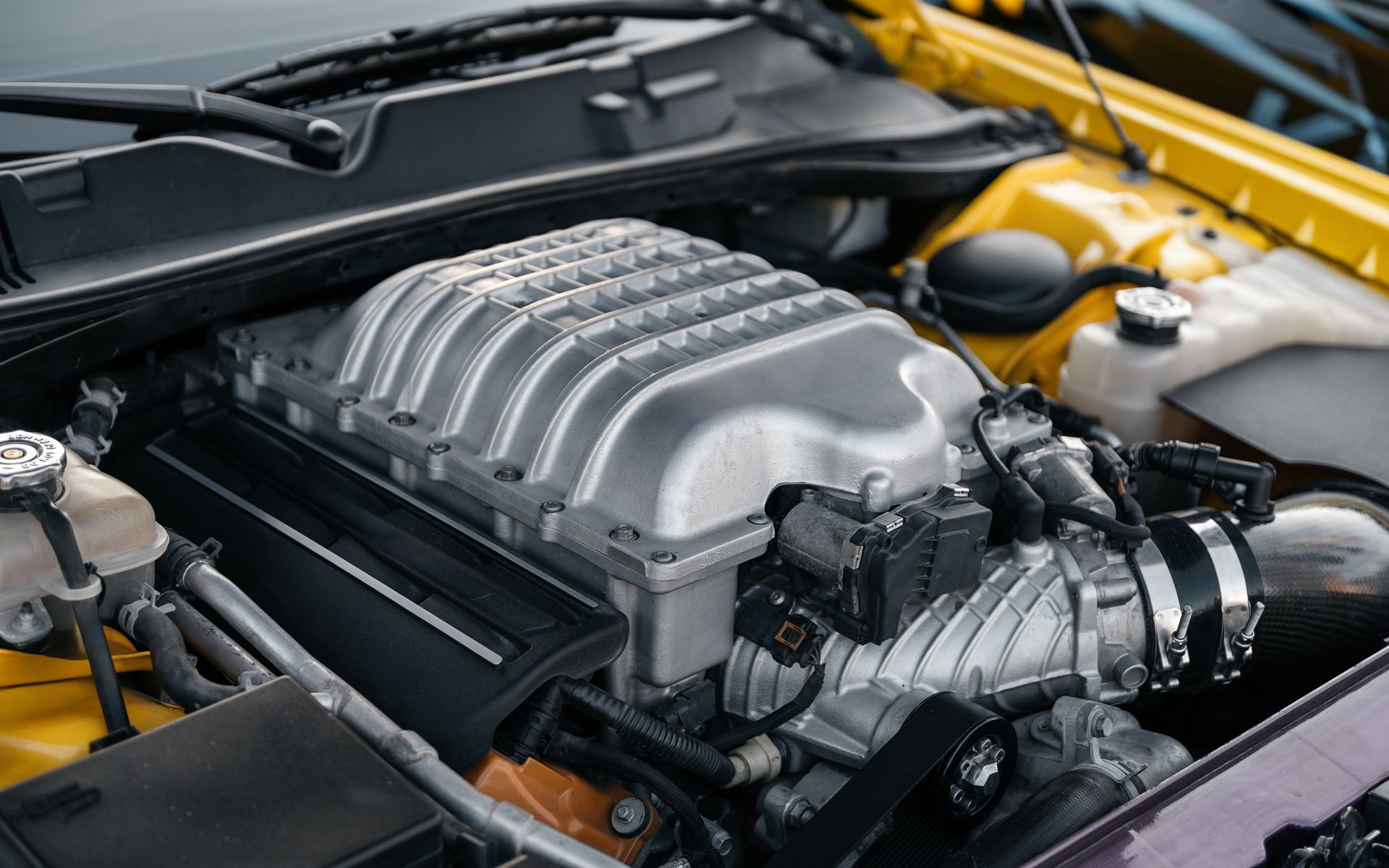 Close-up of a car engine bay with a silver supercharger and black components under the hood