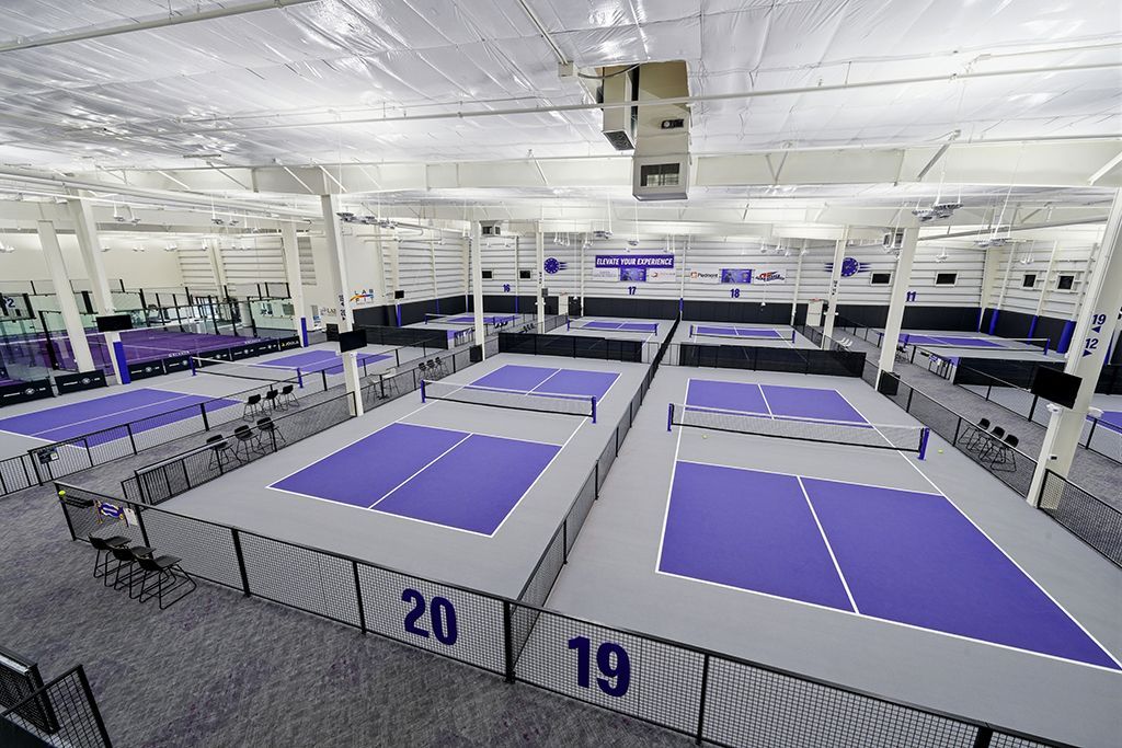 Indoor pickleball courts, gray floors, purple playing areas, metal fences, overhead lighting.