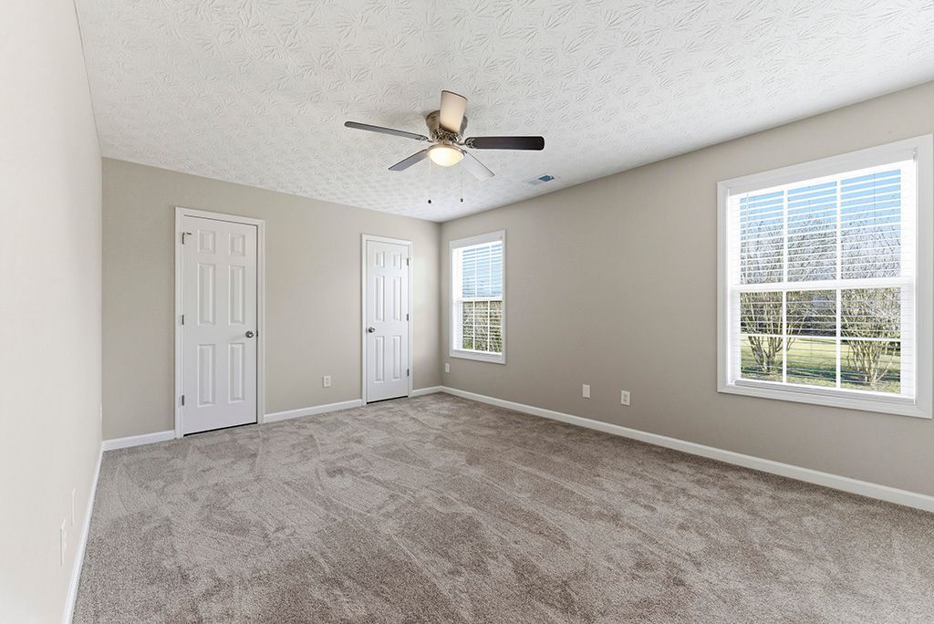 Empty bedroom with beige walls, two doors, two windows, and a ceiling fan.