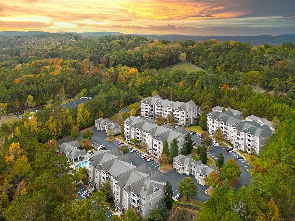 Aerial view of apartment complex nestled in lush, colorful autumnal trees; parking lot, pool visible.