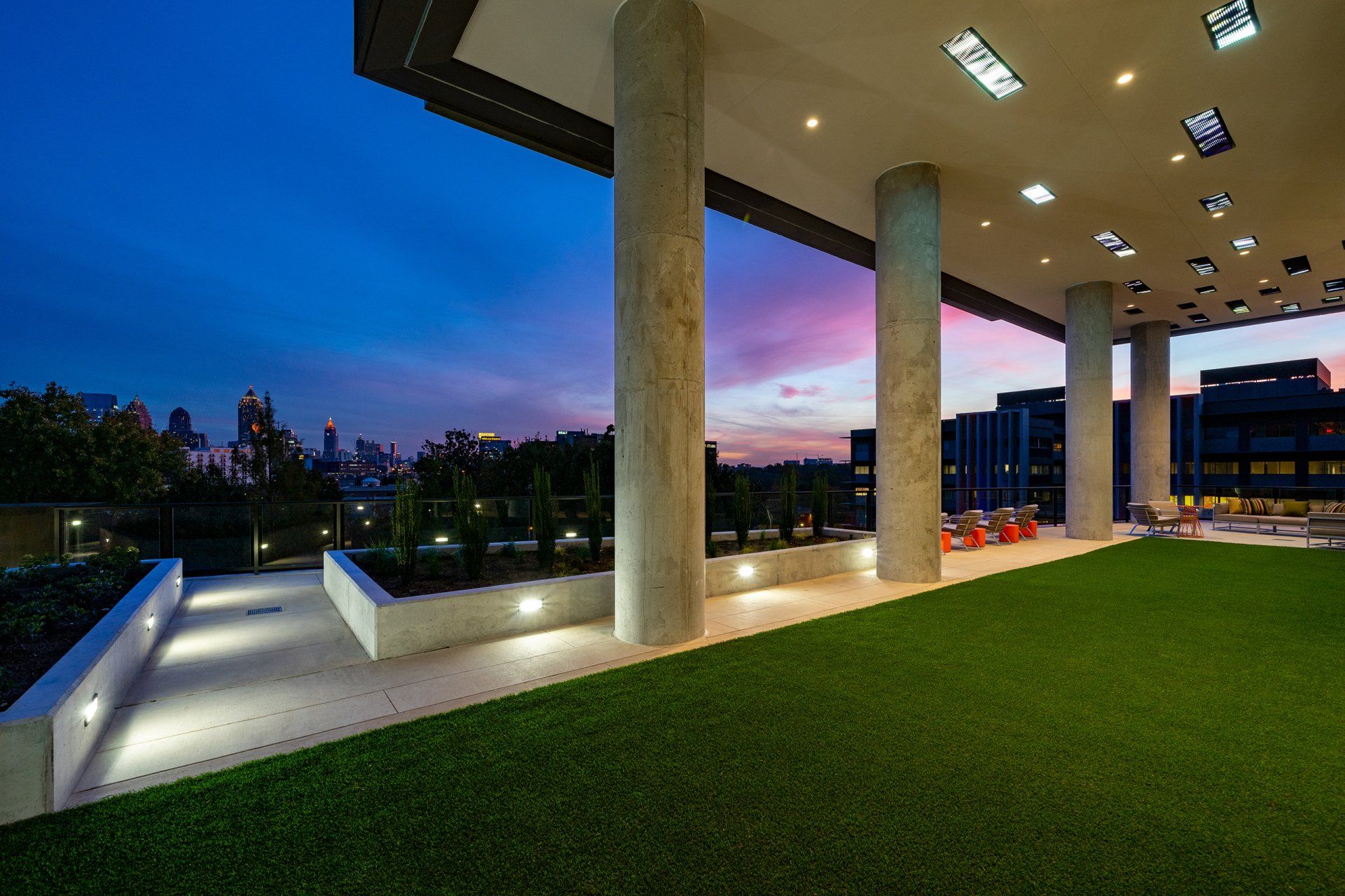 Rooftop patio with green turf, concrete columns, and a city skyline at dusk with pink and blue sky.