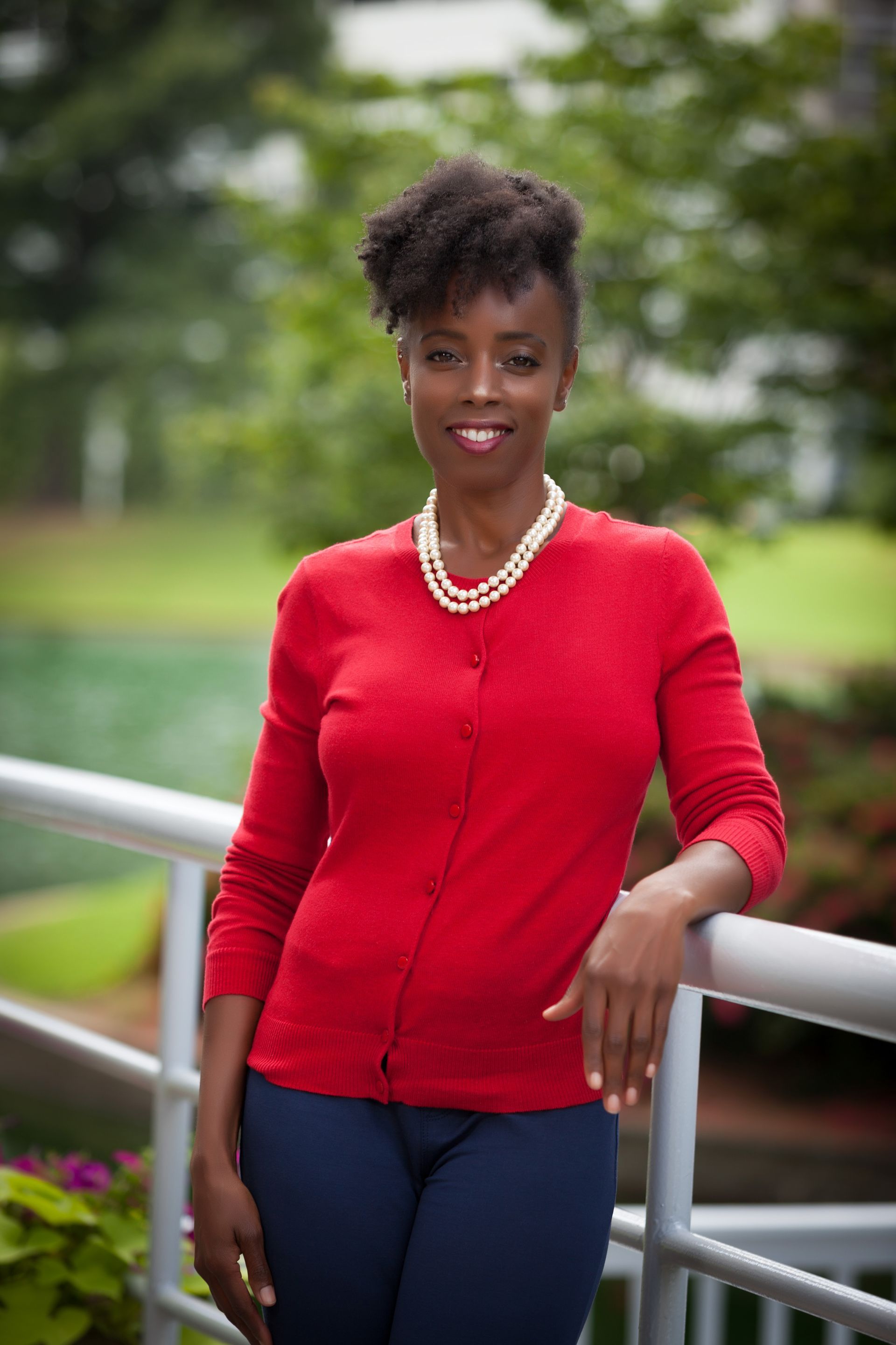 A smiling woman in a red cardigan and pearl necklace stands leaning against a railing in an outdoor setting.