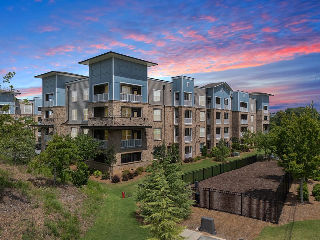 Apartment building with blue accents under a colorful sunset sky. Green landscaping surrounds the building.