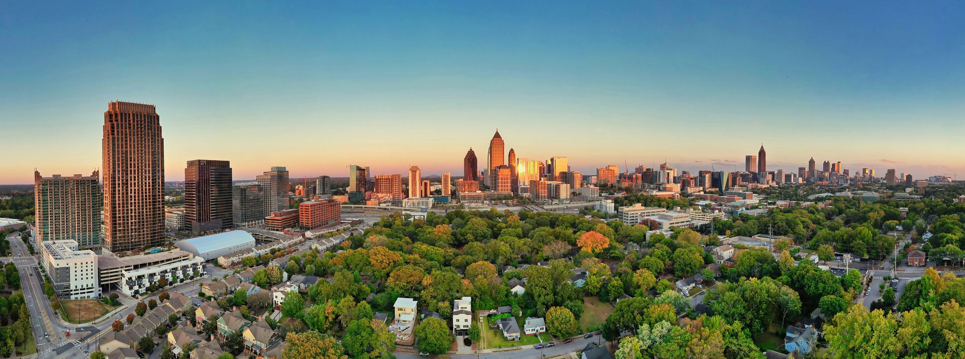  Atlanta skyline at sunset, golden light illuminating buildings. Green park in foreground, blue sky.