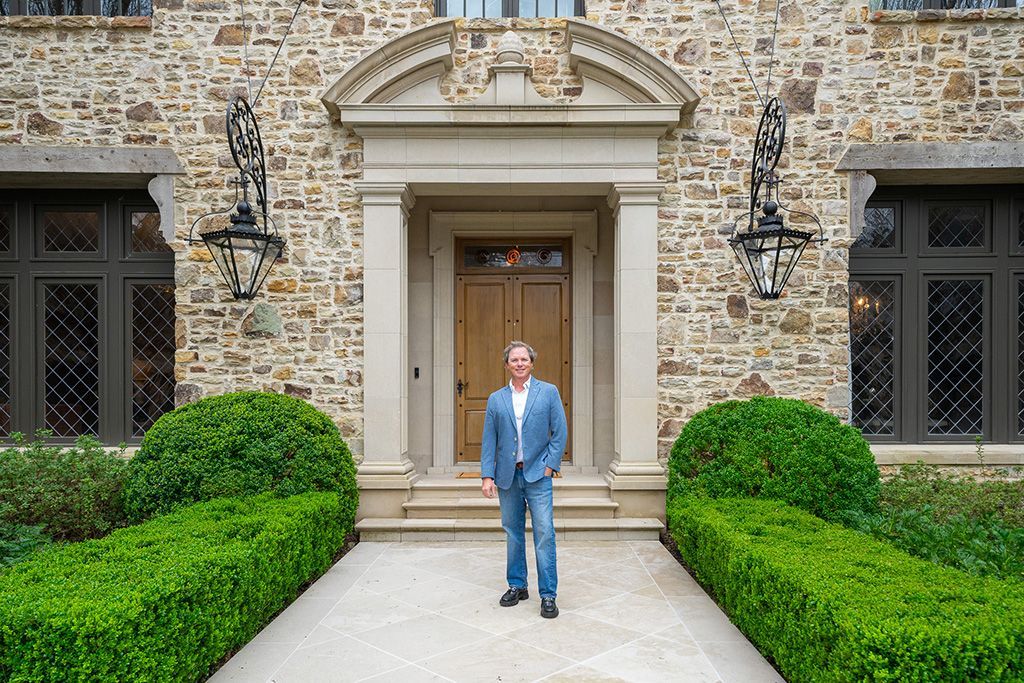 Man in a blue jacket stands at the entrance of a stone house with manicured hedges.
