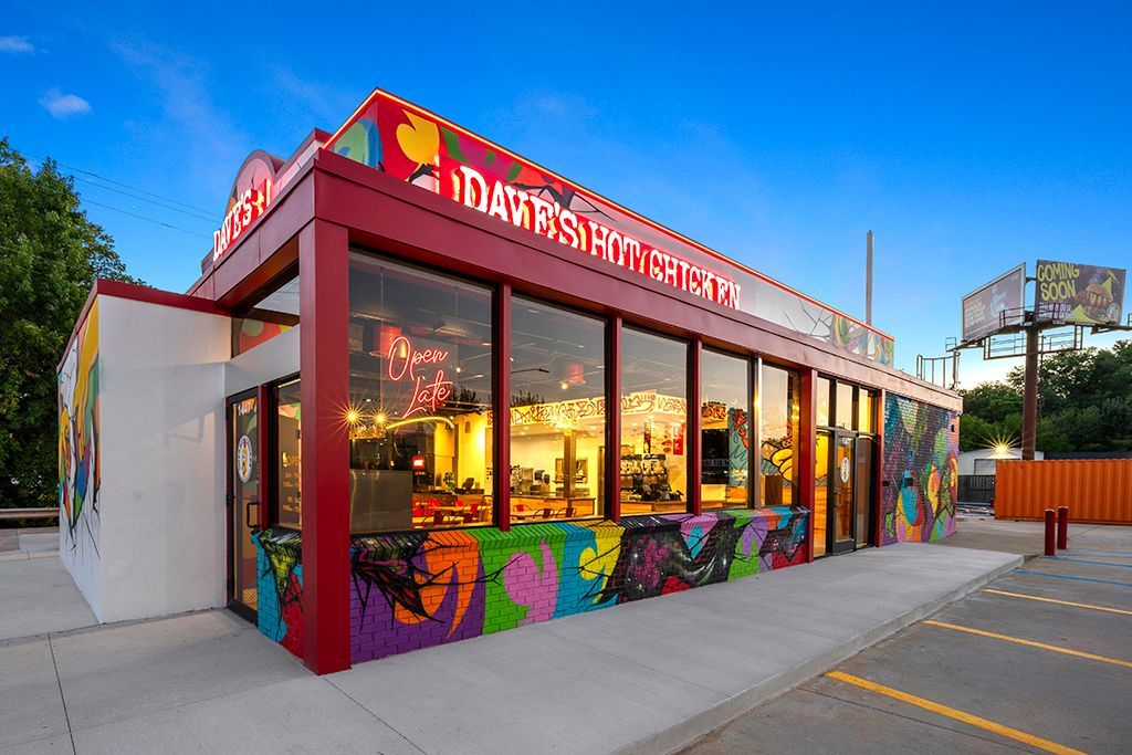 Colorful restaurant with mural-covered walls and large windows under a red awning, parked vehicles in front.