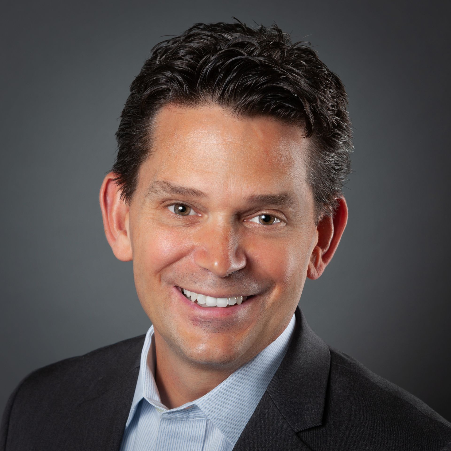 Smiling man in a dark blazer and light blue shirt against a gray studio background
