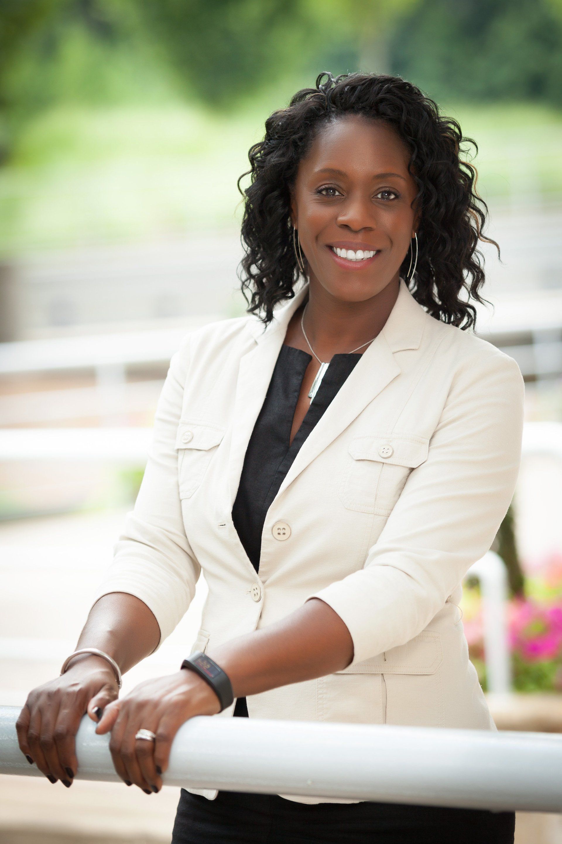 Woman in light blazer, smiling, leaning on railing outside.