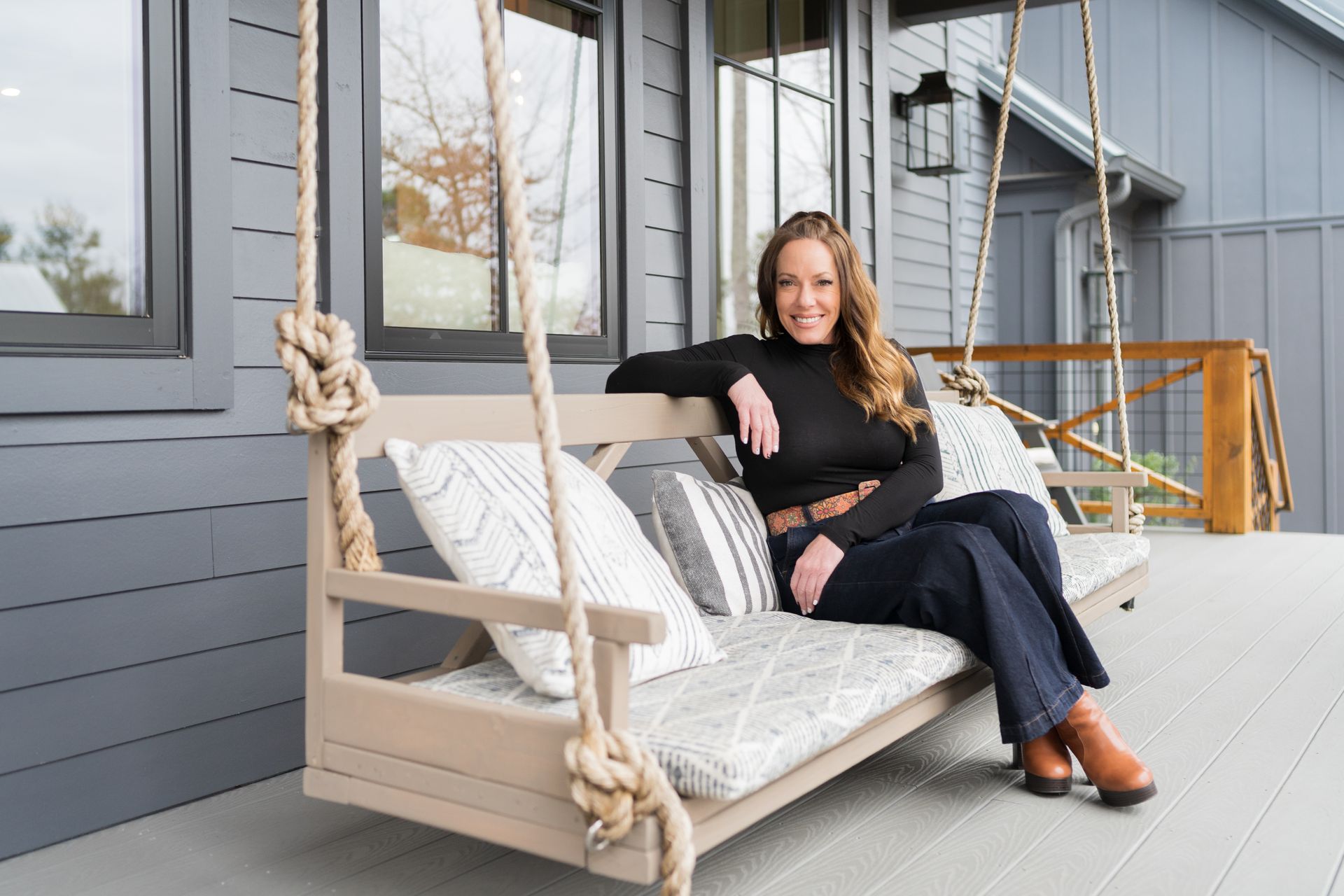 A person sits on a light-colored porch swing on a wooden deck in front of a gray house, smiling at the camera.