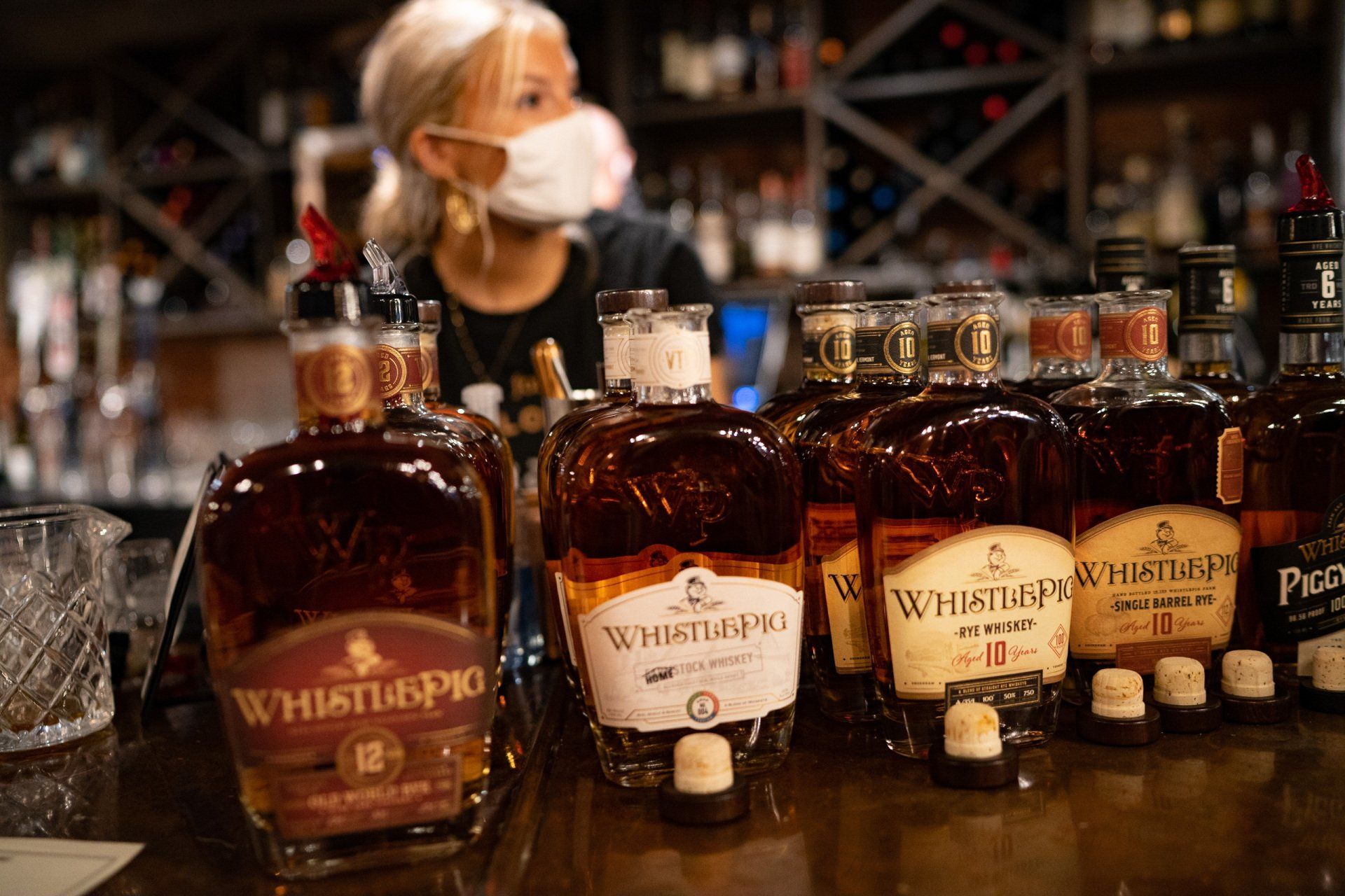 A woman wearing a mask is standing behind a bar filled with bottles of whiskey.