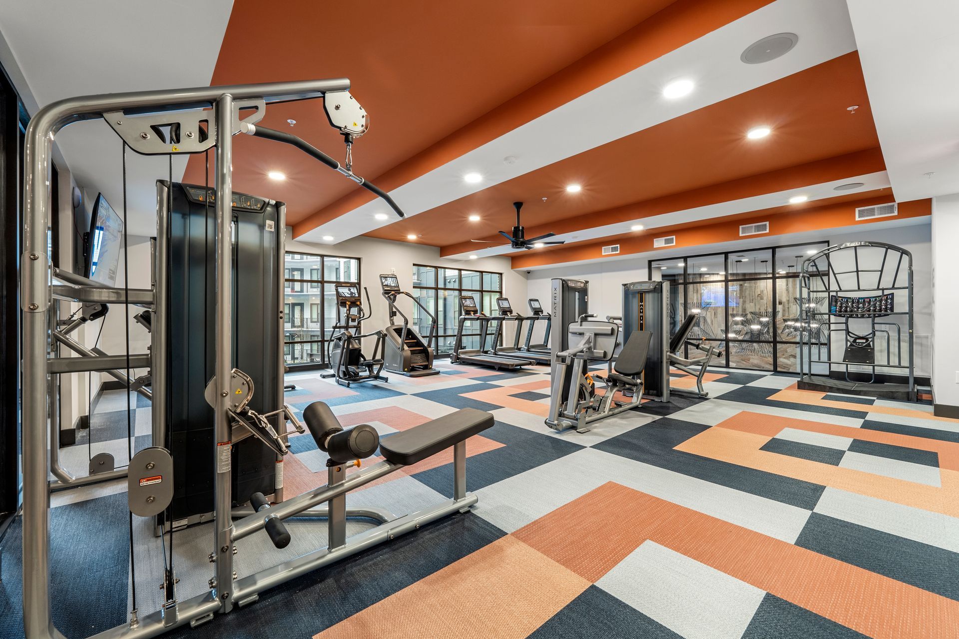 Gym with various exercise machines on patterned floor, orange and white ceiling.