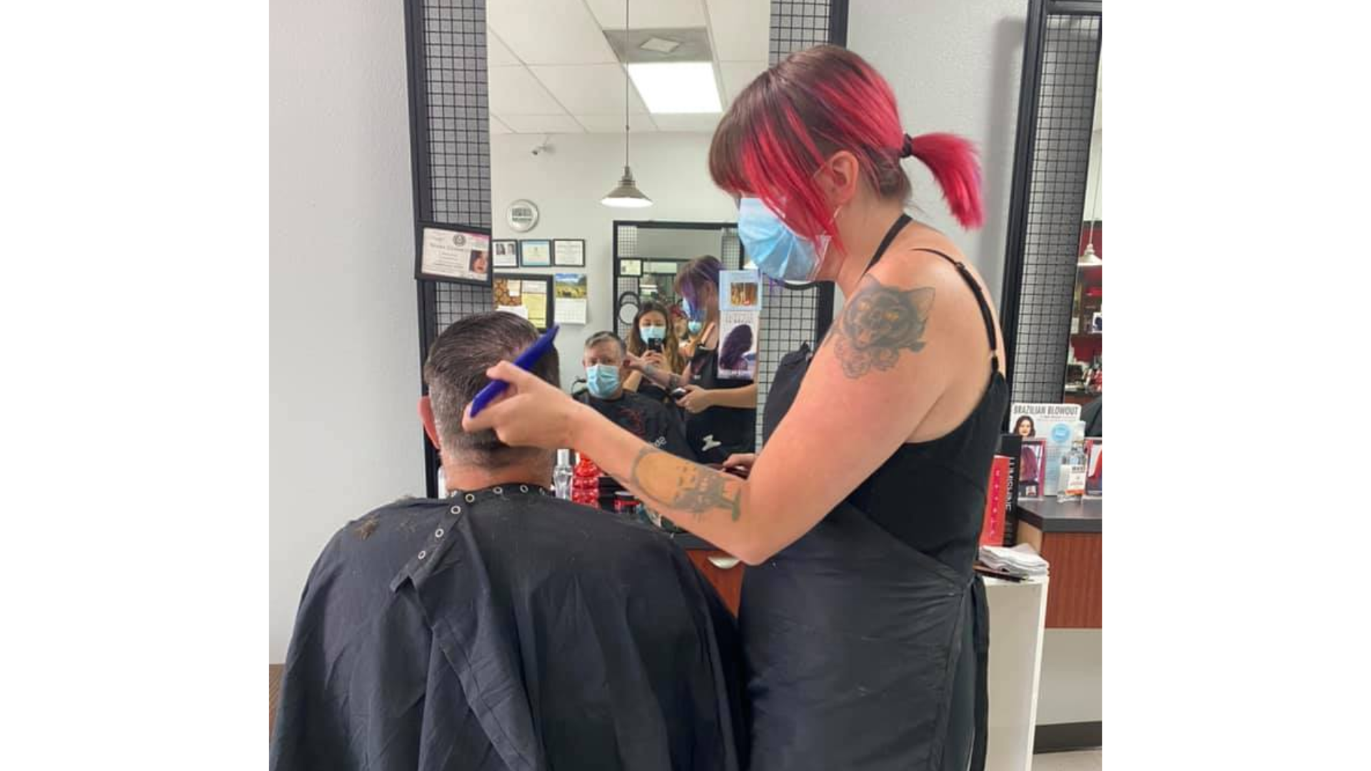 Hairdresser in a salon cutting customer's hair. Both wear masks. Pink hair, arm tattoo, mirrors in background.