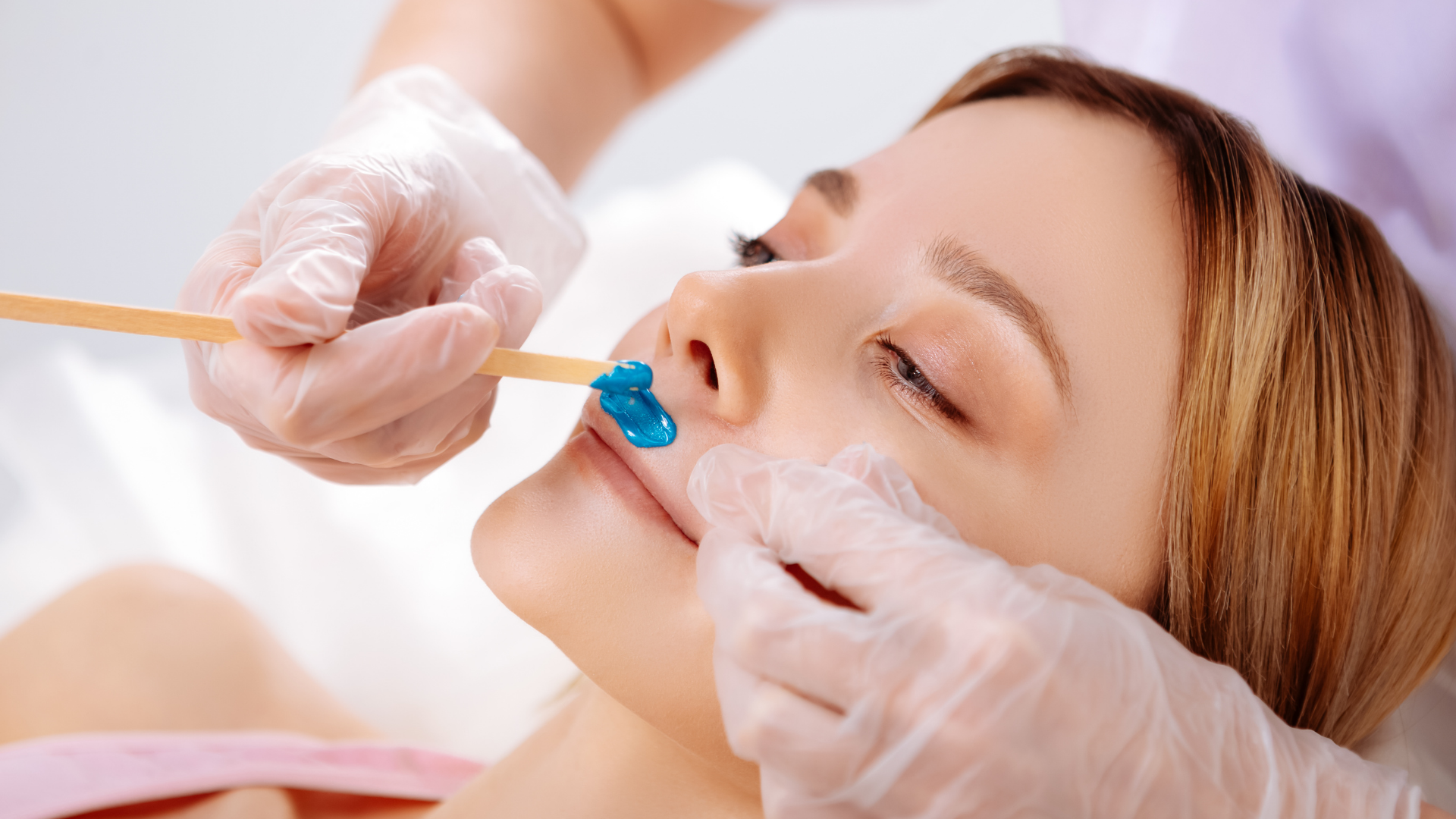 Person getting upper lip waxed at a salon; blue wax applied, gloved hands, wooden applicator, relaxed expression.