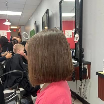 Child with a brown bob haircut in a salon, facing away from the camera. People are in the background, a mirror on the right.