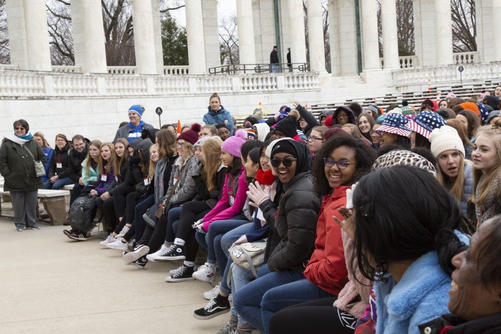 Crowd seated in a long row outdoors near a white stone building, facing left