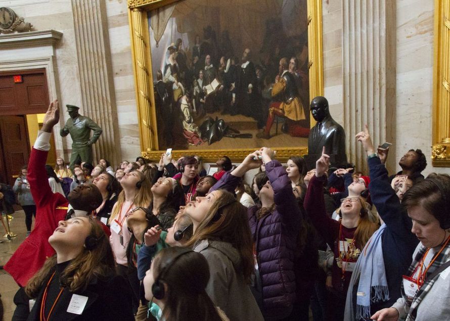 Crowd in an ornate hall raising hands toward a statue and large painting on the wall