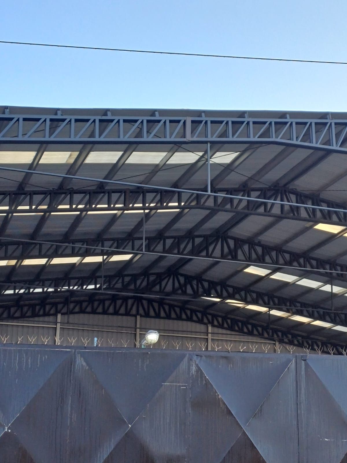 Estructura de techo de cerchas de acero contra un cielo azul claro, con una valla gris en primer plano.