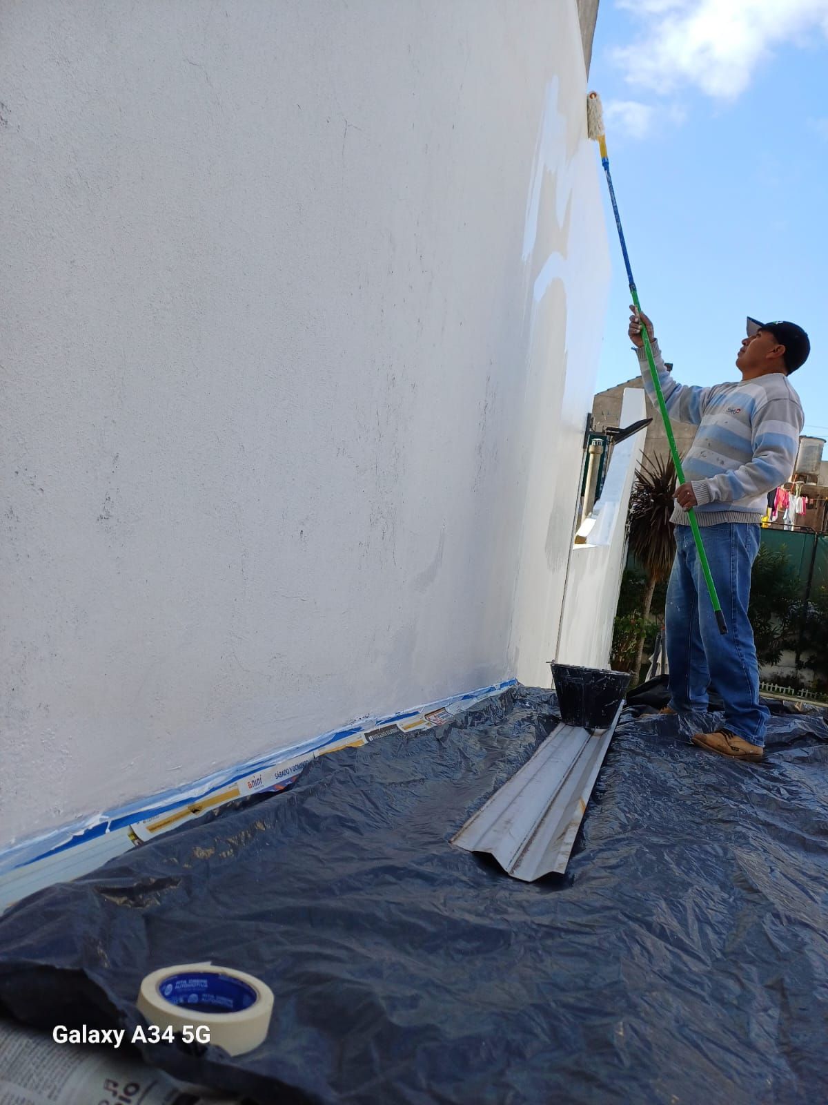 Una persona pinta una pared exterior blanca con un rodillo junto a un suelo cubierto con una lona negra.