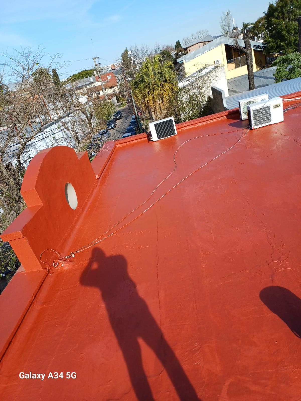 Azotea roja con rejillas de ventilación y sombra, con vistas a árboles y casas en un día soleado.