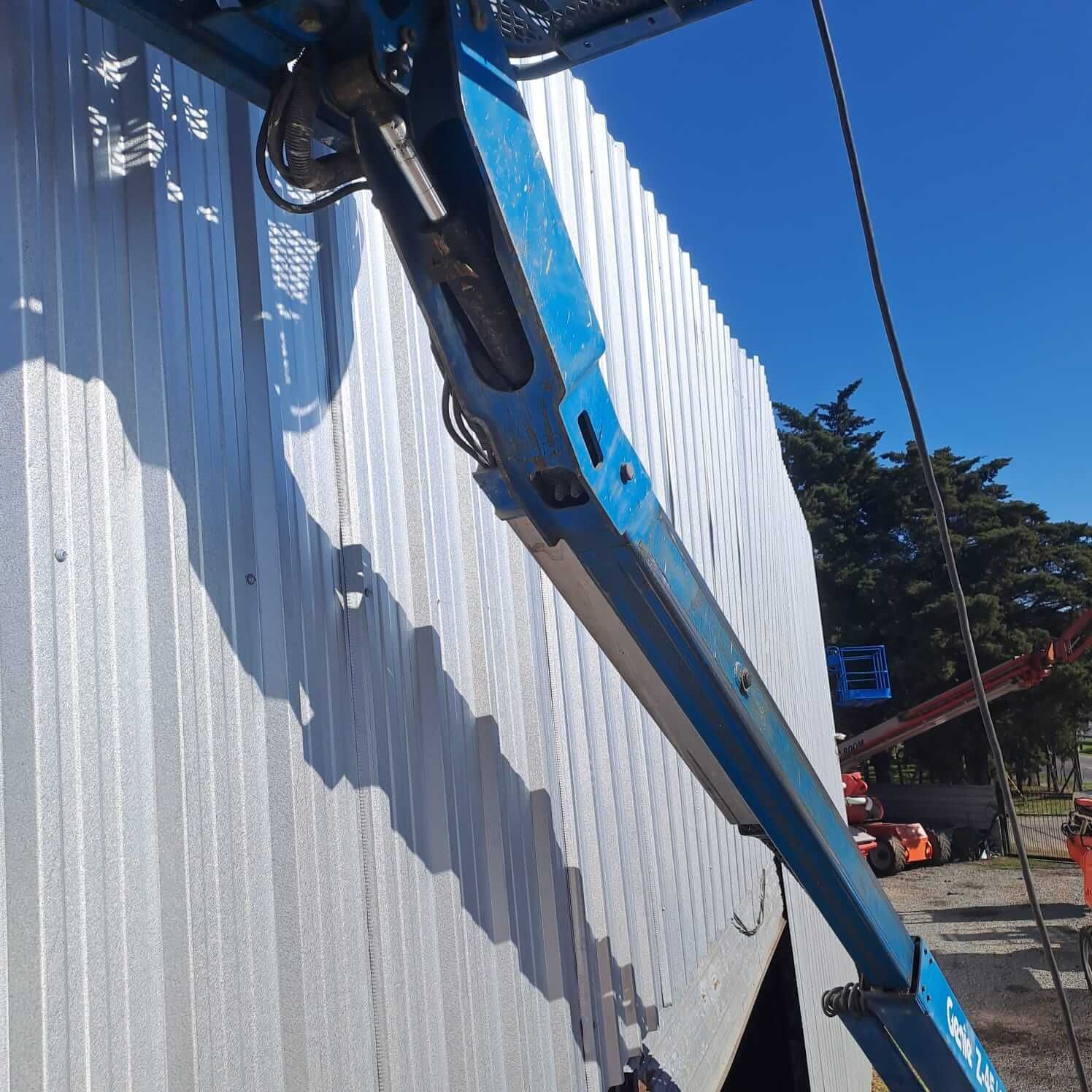 Una plataforma elevadora azul trabaja junto a una pared blanca ondulada bajo un cielo despejado.