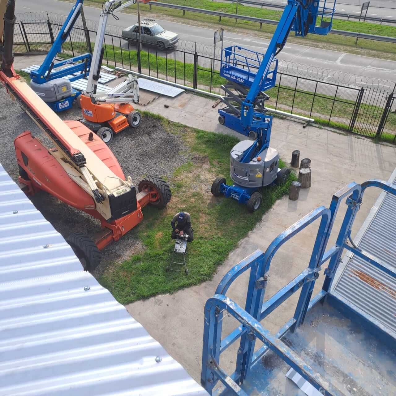 Obra en construcción con elevadores azules y naranjas, un trabajador abajo y una carretera al fondo.