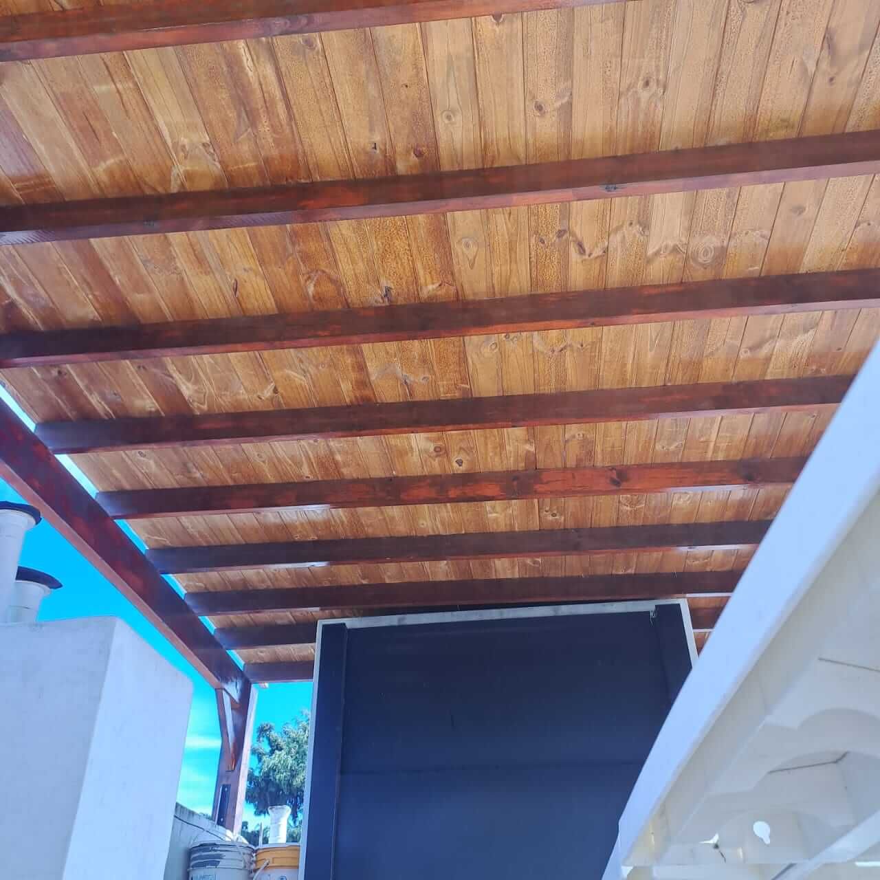 Techo de listones de madera sobre un patio con una puerta oscura y una pared blanca debajo.
