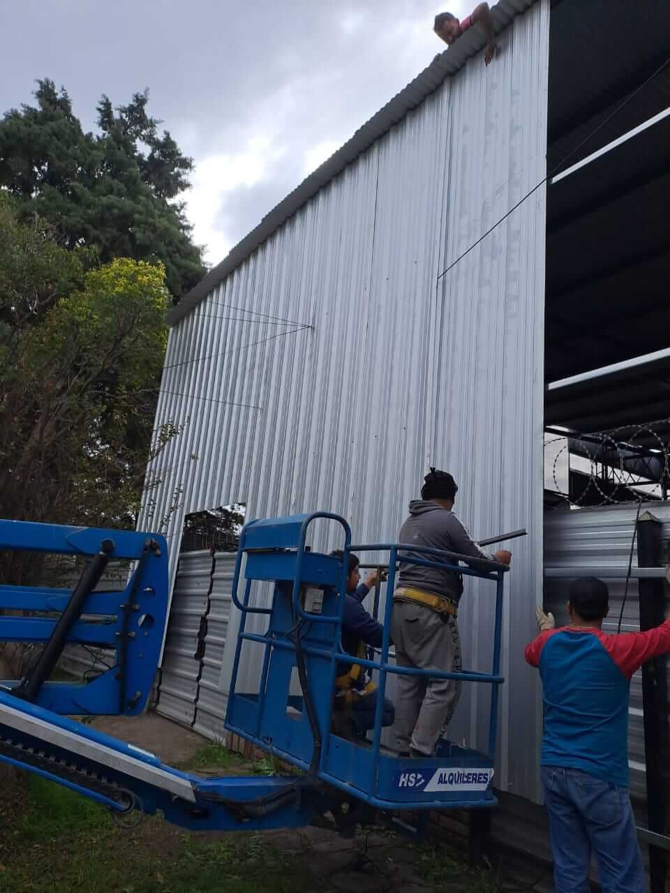 Trabajadores en plataformas elevadoras azules reparan el exterior de un gran edificio metálico blanco.