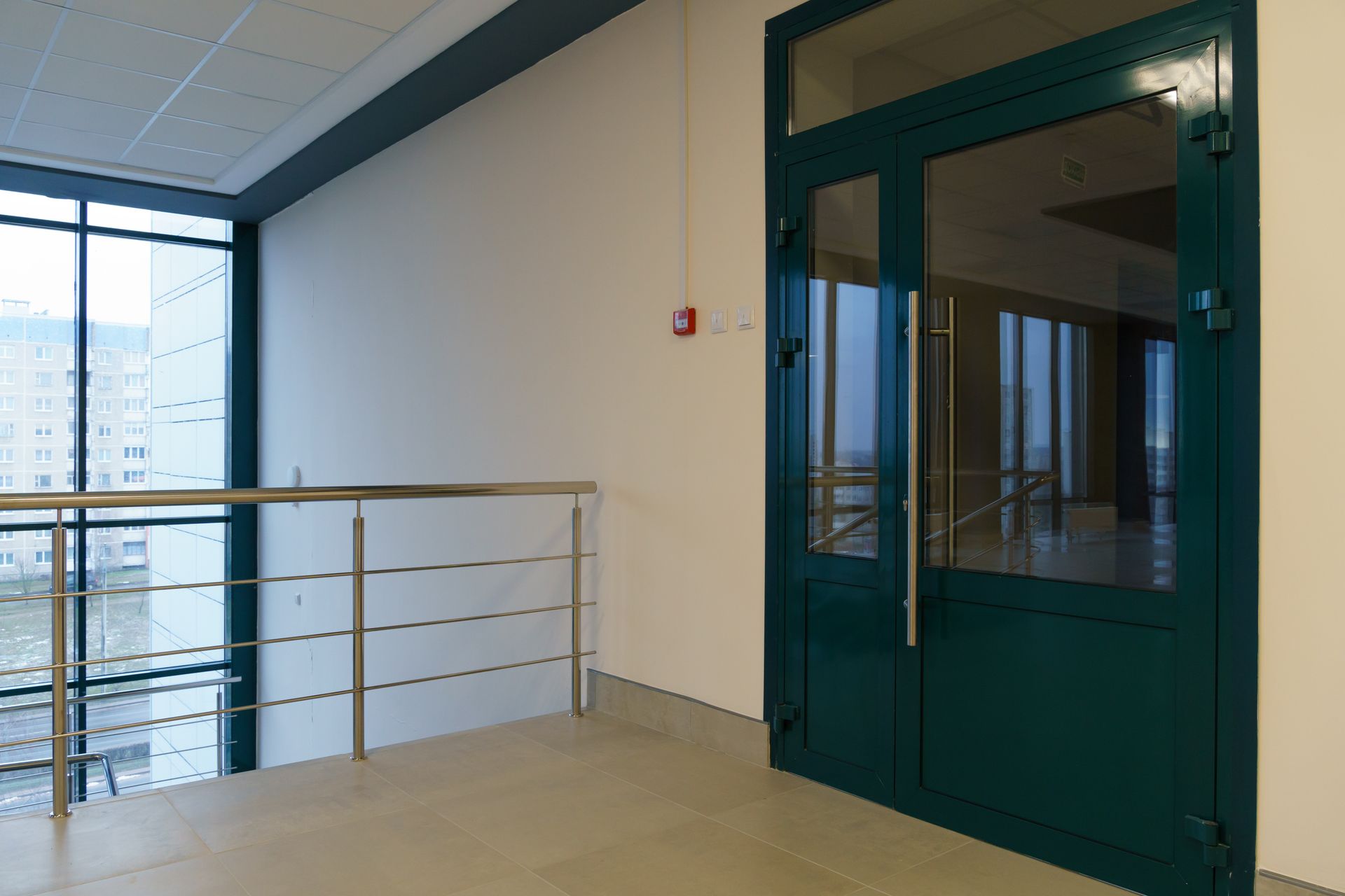 Interior hallway with teal door, railing, and large window overlooking a building exterior