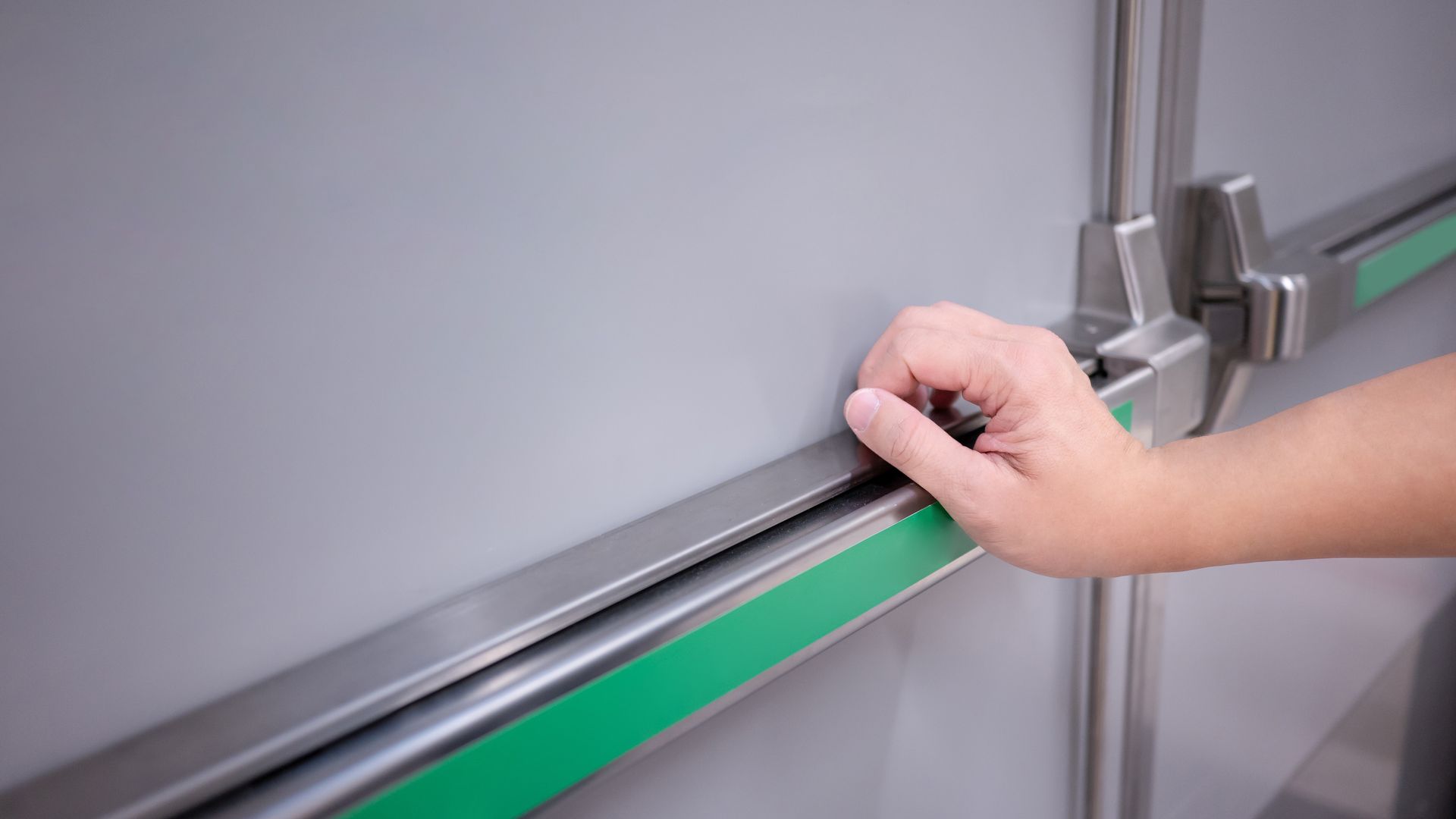 Hand presses the bar on a grey emergency exit door with a green stripe.