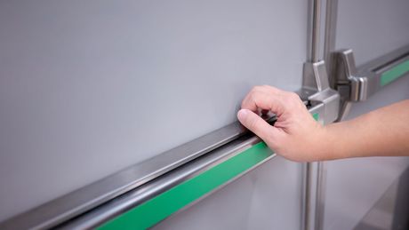 Hand presses the bar on a grey emergency exit door with a green stripe.