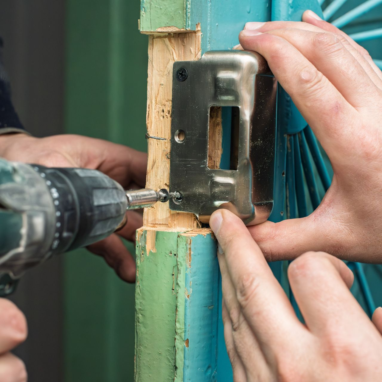 Hands installing a door latch with a power drill on a turquoise door frame.