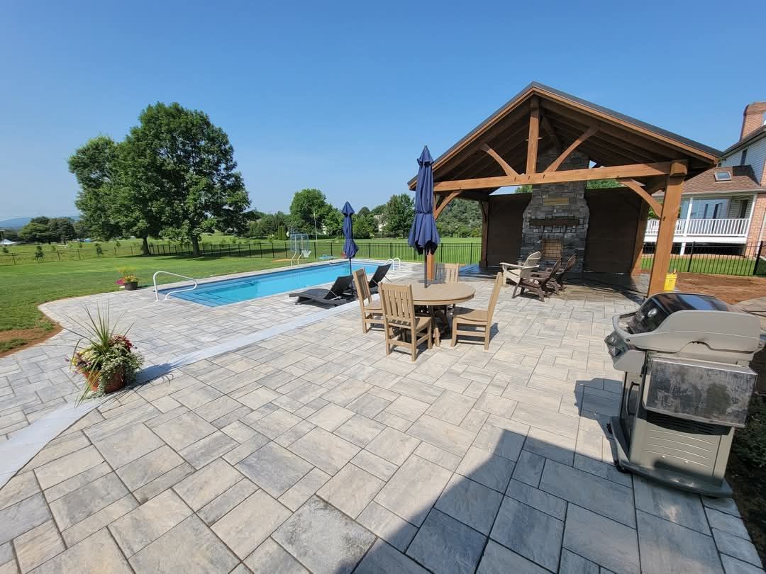 Jesse James Hardscaping - Patio with pool, dining set, grill, and stone fireplace under a wood-beamed pavilion on a sunny day.