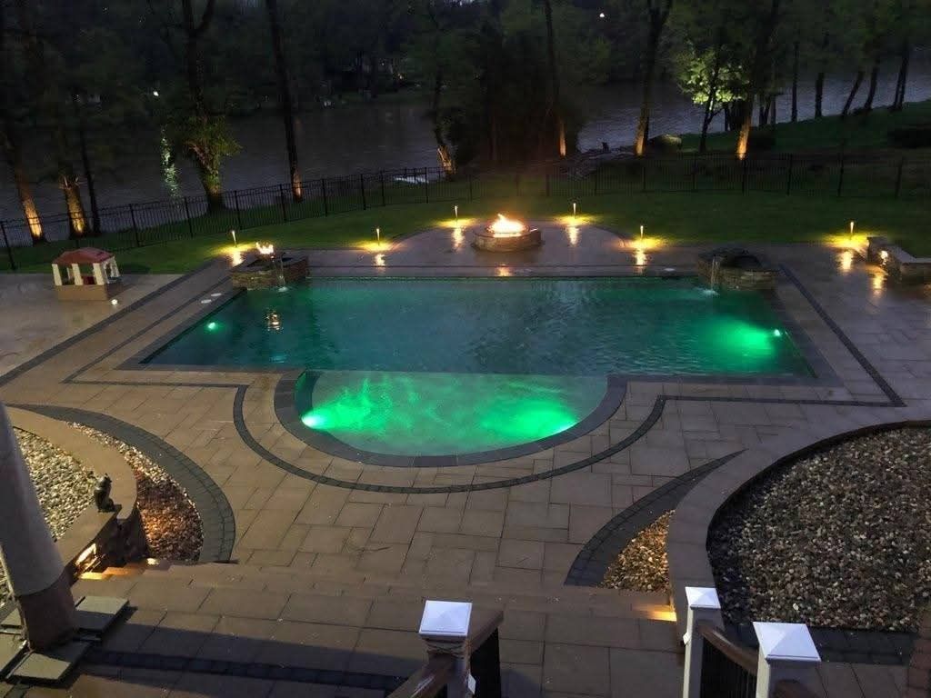Jesse James Hardscaping - Nighttime view of a pool with green lights, a fire pit, and surrounding patio with trees and a river in the background.