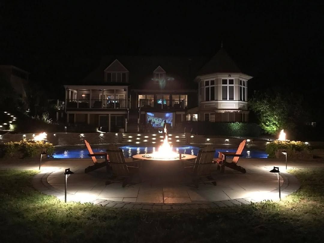 Jesse James Hardscaping - Nighttime view of a house with a fire pit, pool, and outdoor lighting. Two chairs face the fire.