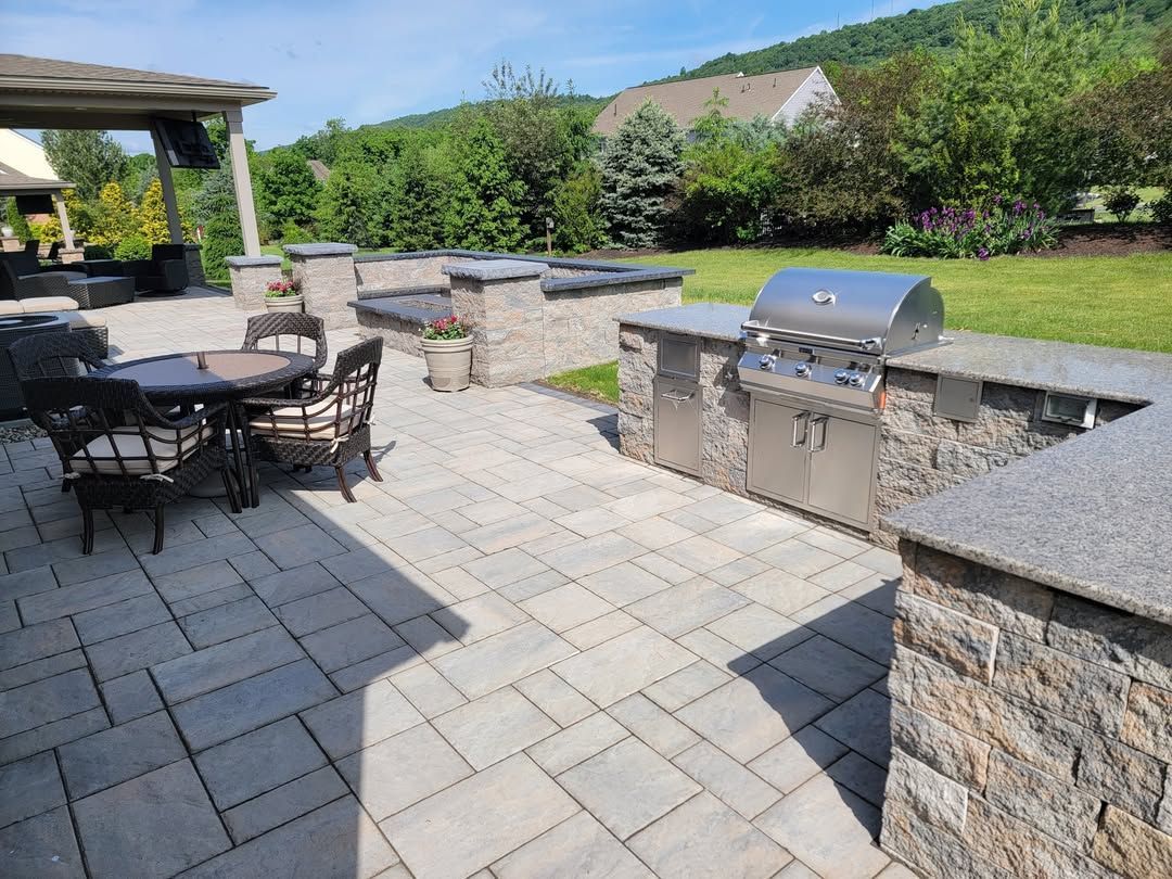 Jesse James Hardscaping - Image -Outdoor patio with grill, seating, and stone walls. Sunny day with grass and trees in the background.