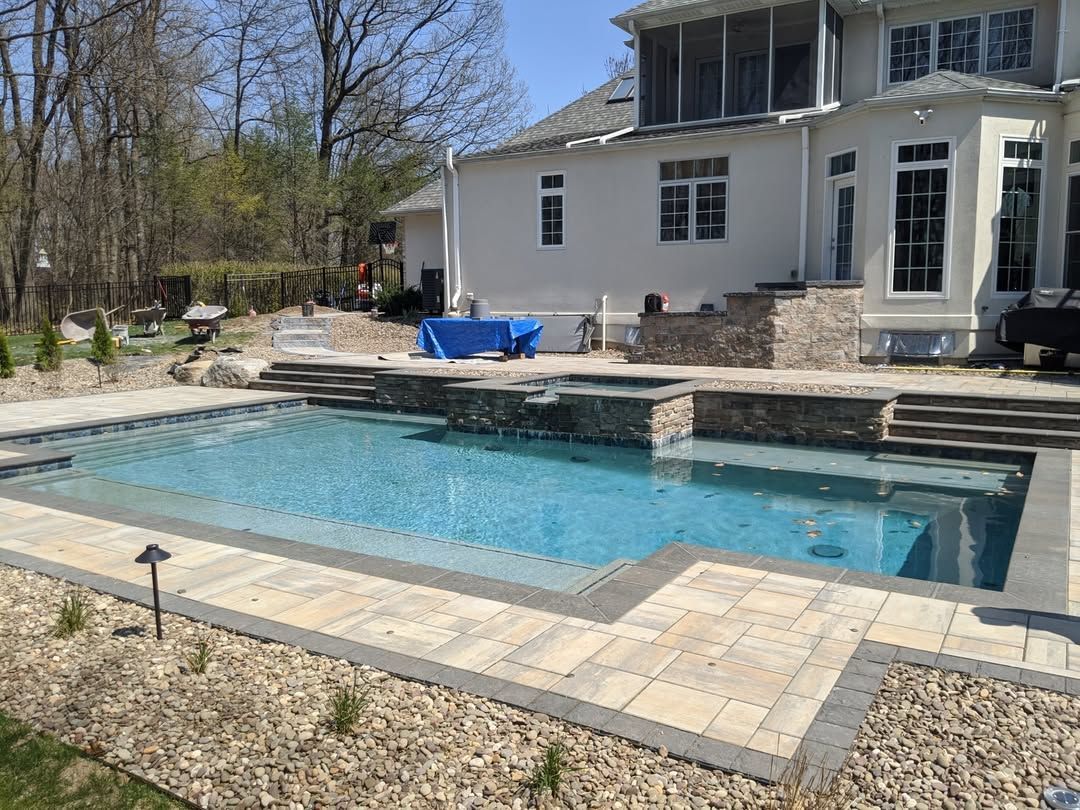 Jesse James Hardscaping - Image -Swimming pool with stone deck and landscaping beside a large house on a sunny day.