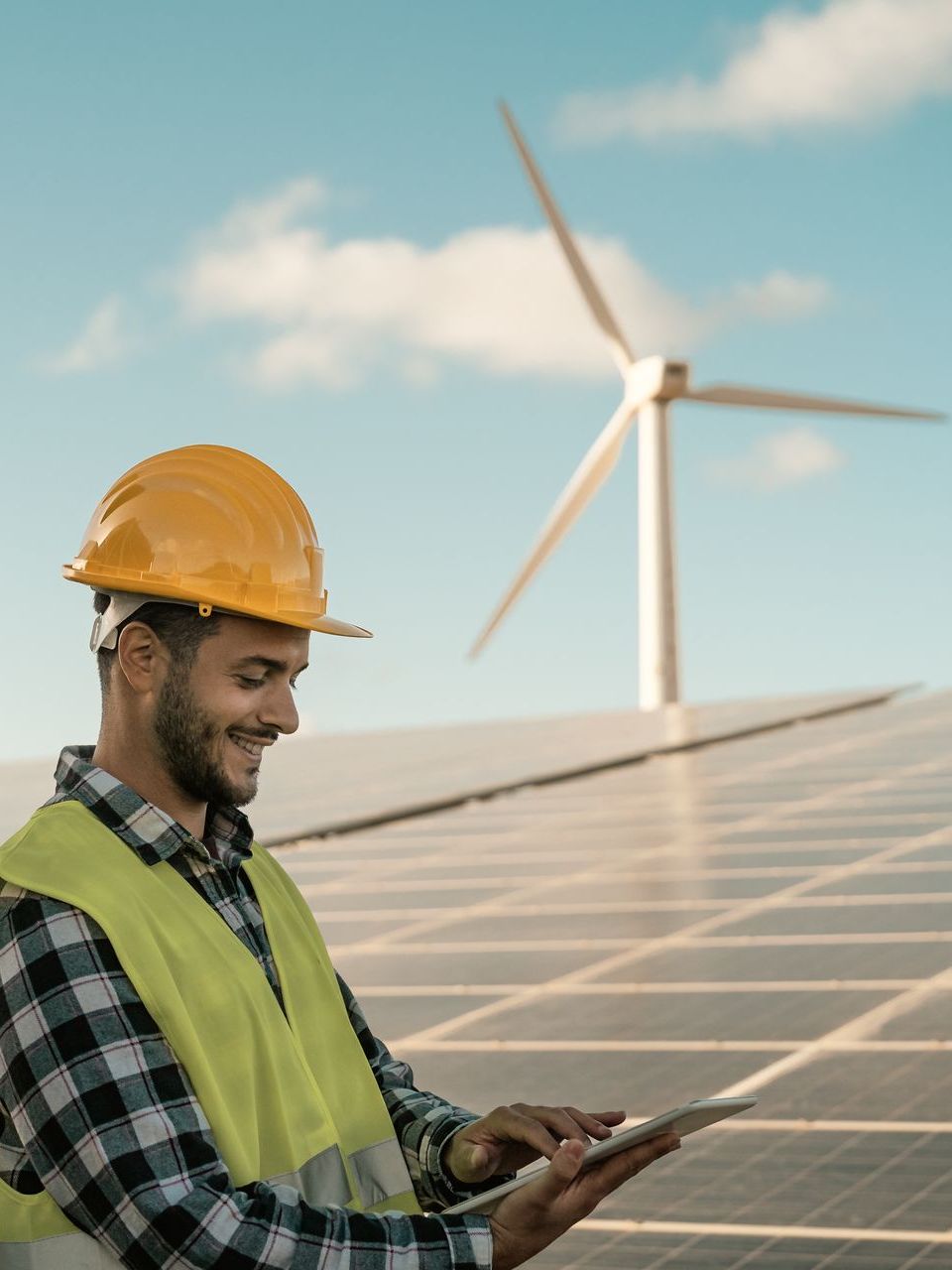 ein Mann mit Helm und Weste schaut auf ein Tablet vor einer Windturbine