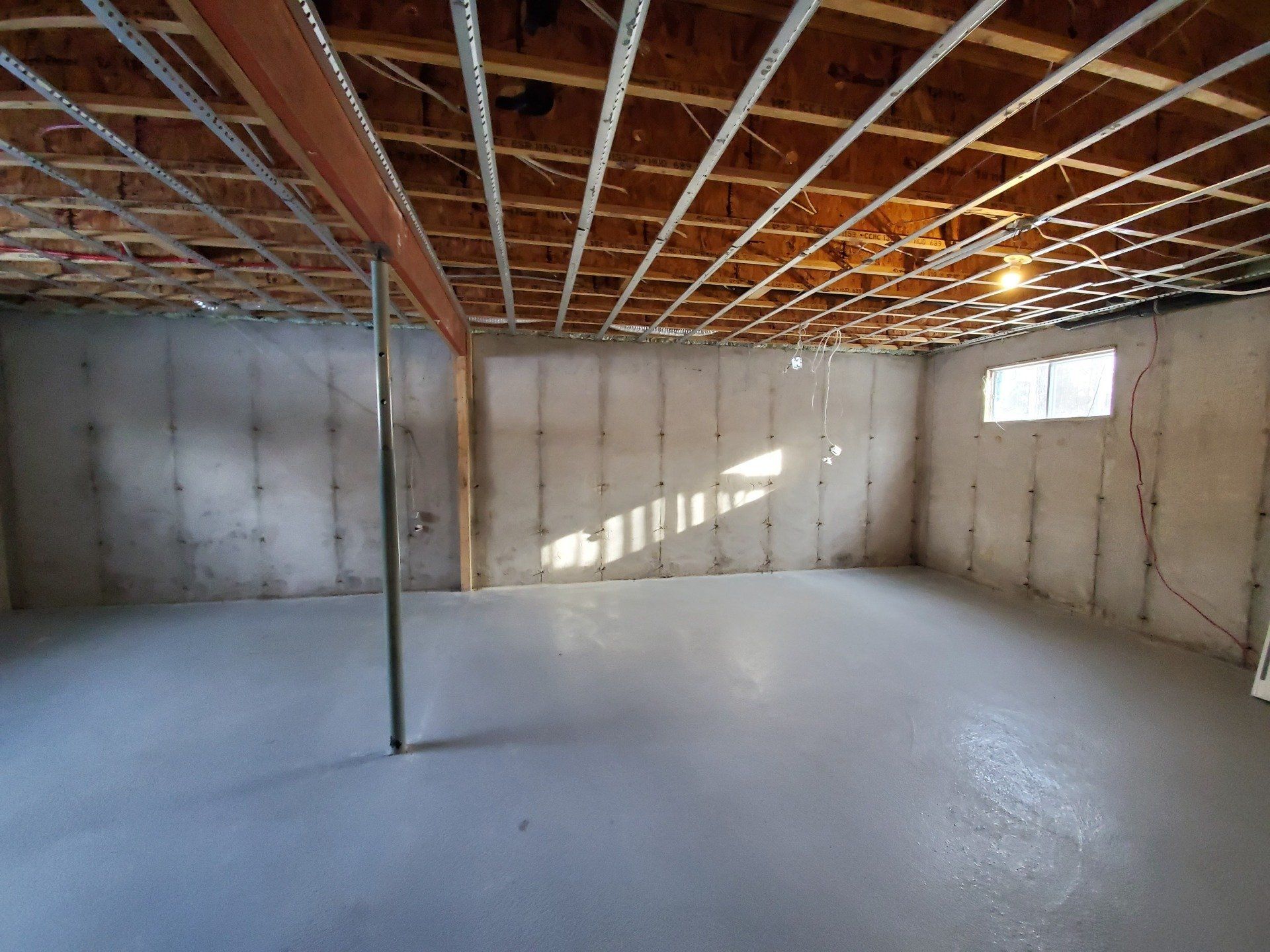 Sous-sol non fini avec murs et plancher en béton, solives de plafond apparentes, poteau de support et une petite fenêtre.