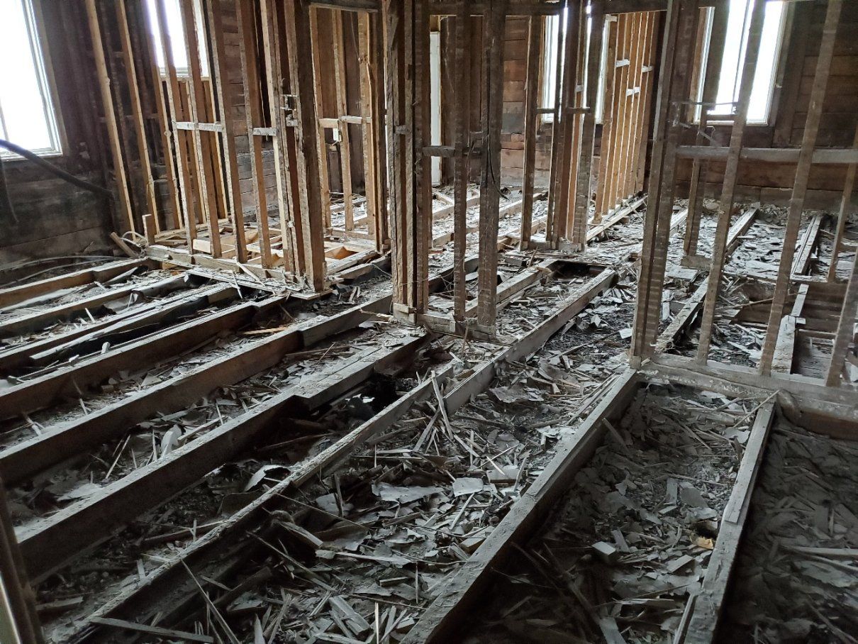 Pièce intérieure en cours de démolition, exposant la charpente en bois et les solives de plancher, débris éparpillés.